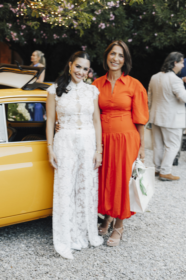The bride stands with a guest in an orange dress beside the yellow car.