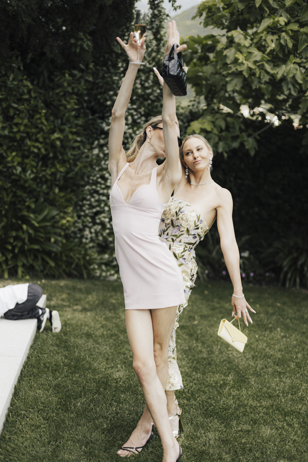 Two guests dance with raised arms in the garden, one holding a glass.
