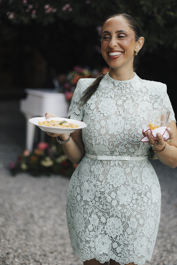 A guest in a pale lace dress smiles while carrying small plates of food.