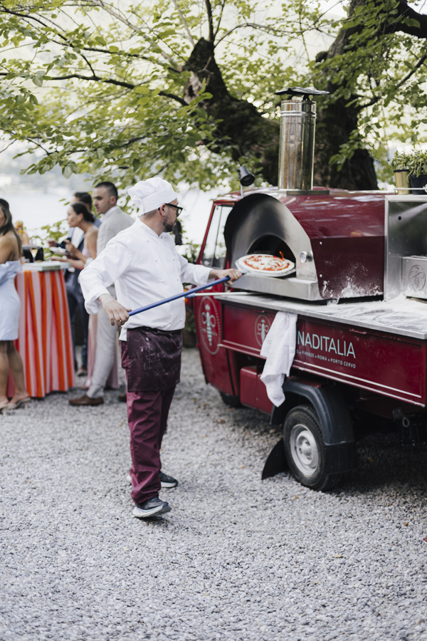 A chef tends the red pizza oven while guests gather in the garden.