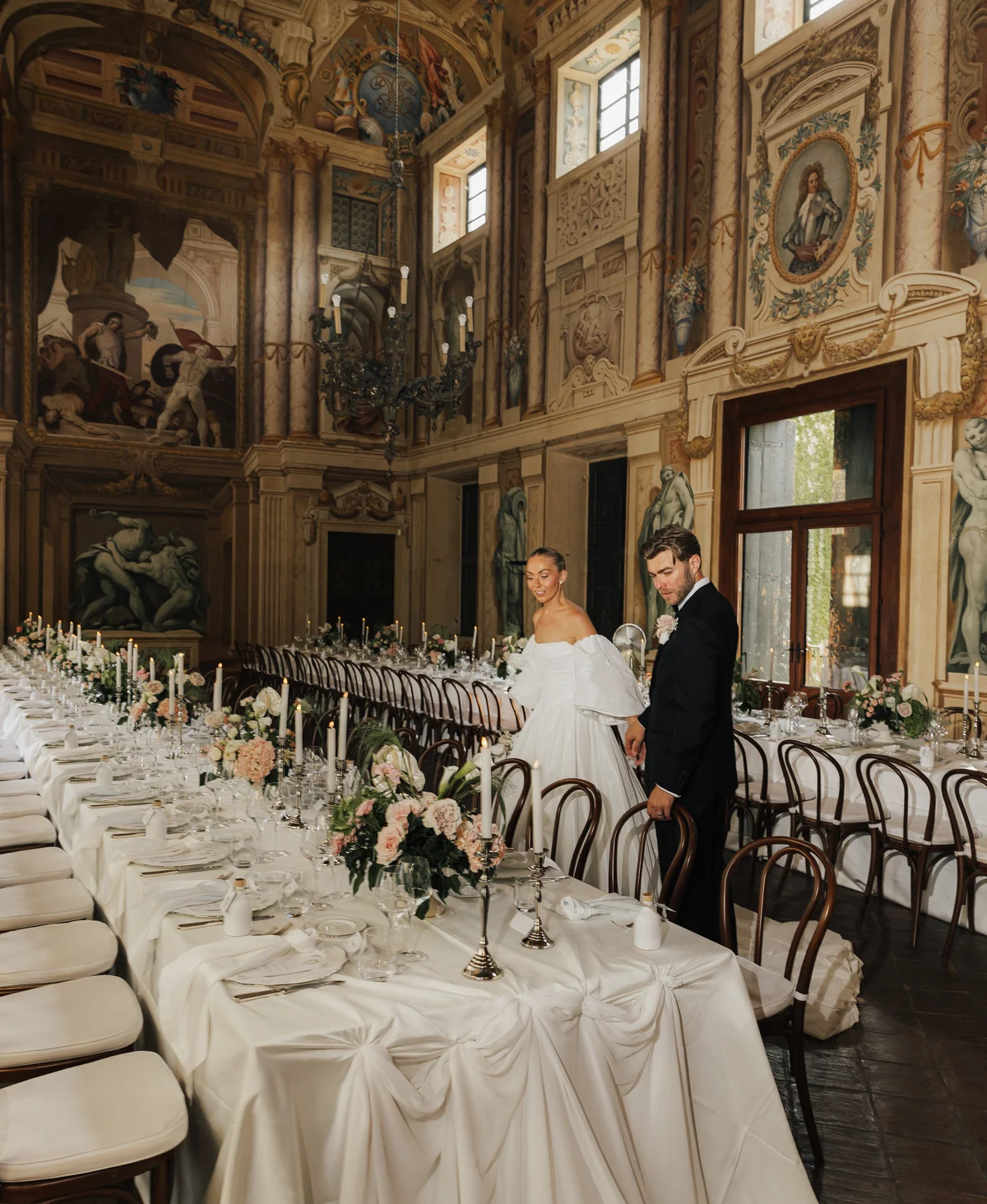 Newlyweds beside a candlelit reception table in a frescoed hall